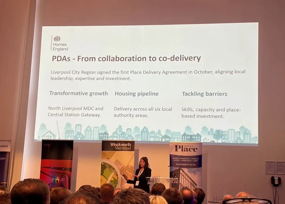 A speaker at Place RESI 2026 presenting a Homes England slide titled “PDAs: from collaboration to co-delivery”. It says Liverpool City Region signed the first Place Delivery Agreement in October, aligning local leadership, expertise and investment, under three headings: transformative growth (North Liverpool MDC and Central Station Gateway), housing pipeline (delivery across all six local authority areas) and tackling barriers (skills, capacity and place-based investment).